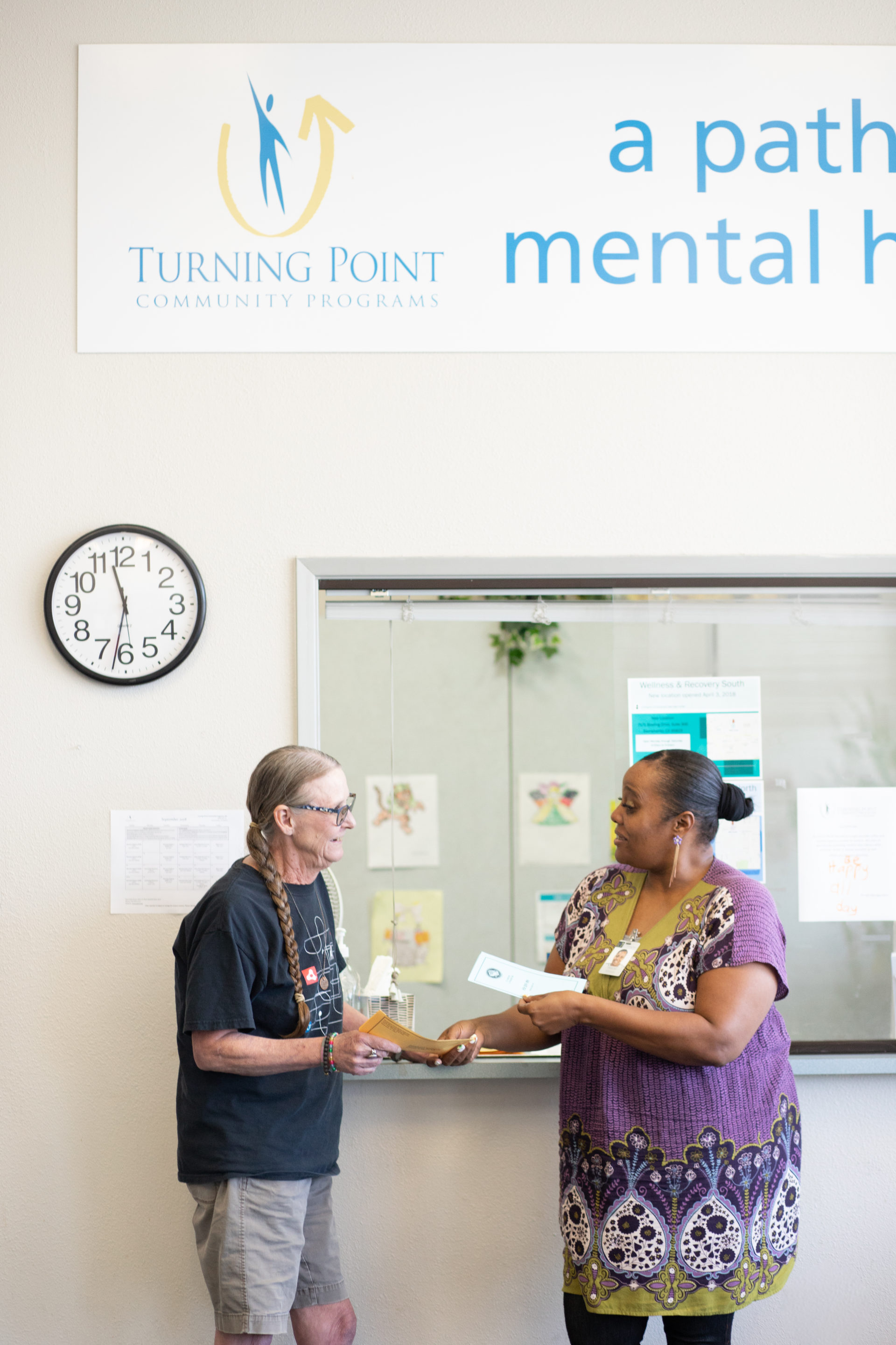 Photo of a woman handing brochures to another person in front of a recption window, under a banner with the Turning Point Community Programs logo on it.