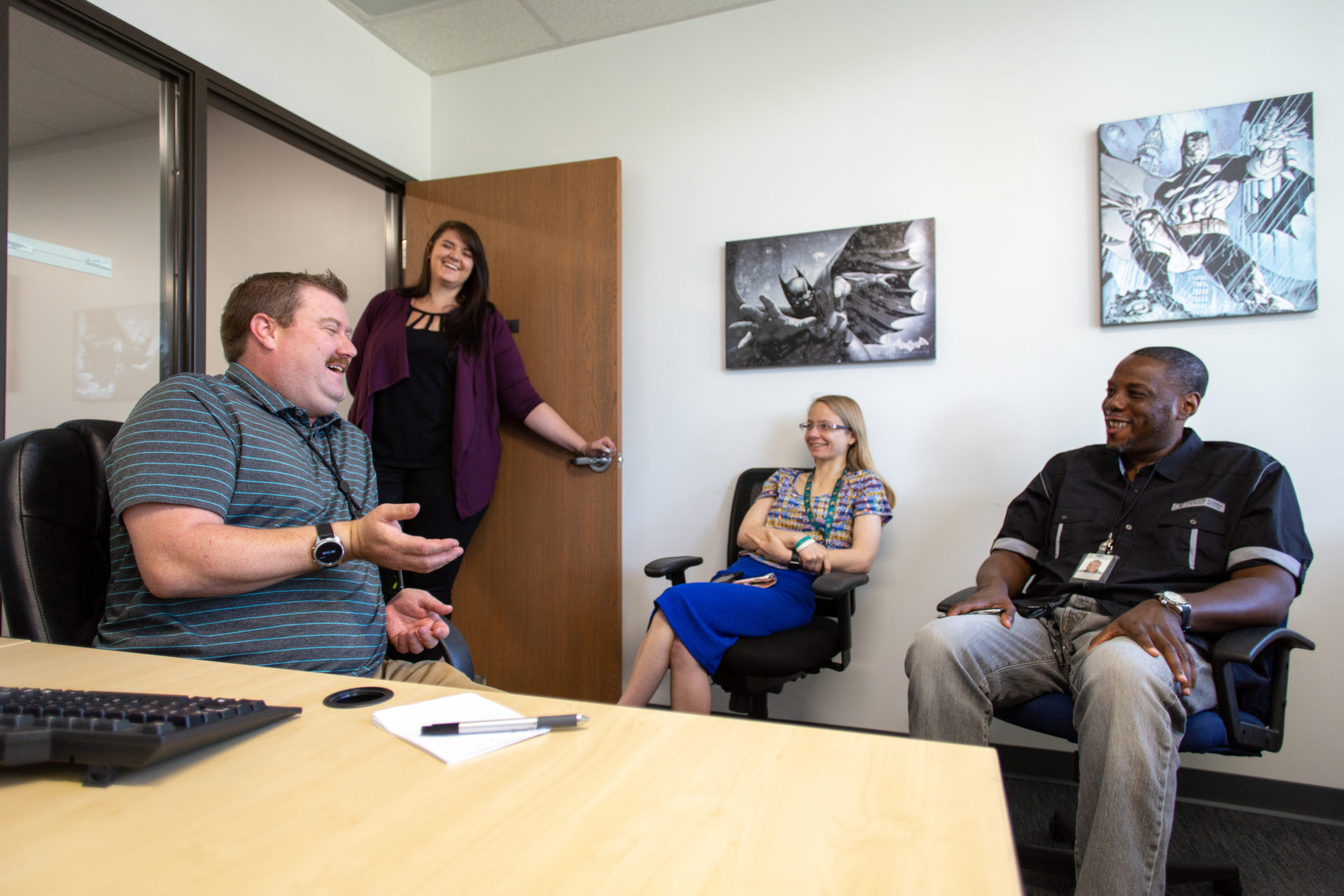 Photo of group of employees sitting in office talking happily as a smiling woman enters the room to join them.